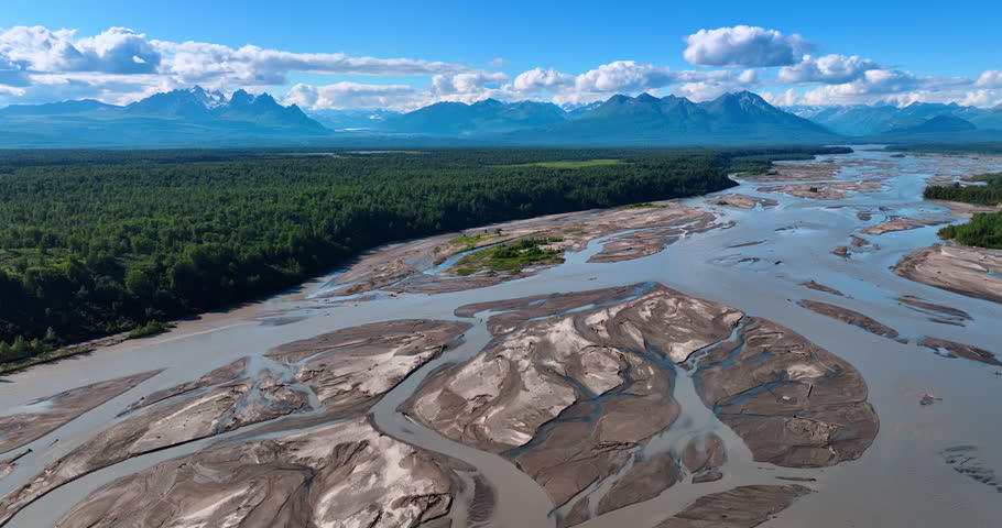 Delta of the wide shallow river surrounded by the pine tree woods. Mountain range at backdrop. Flight over the wild nature of Alaska.