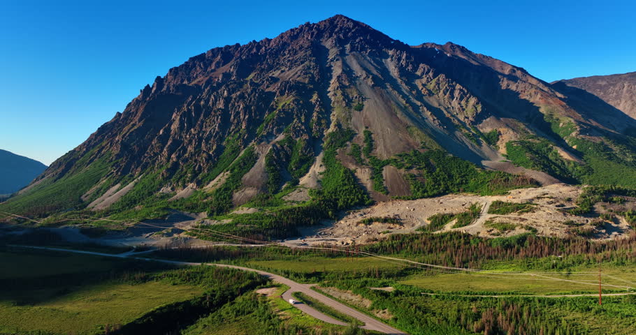 Huge bare rock with scarce vegetation on the slopes. Footage above the valley crossed by the highway and electricity line. Alaska landscapes.