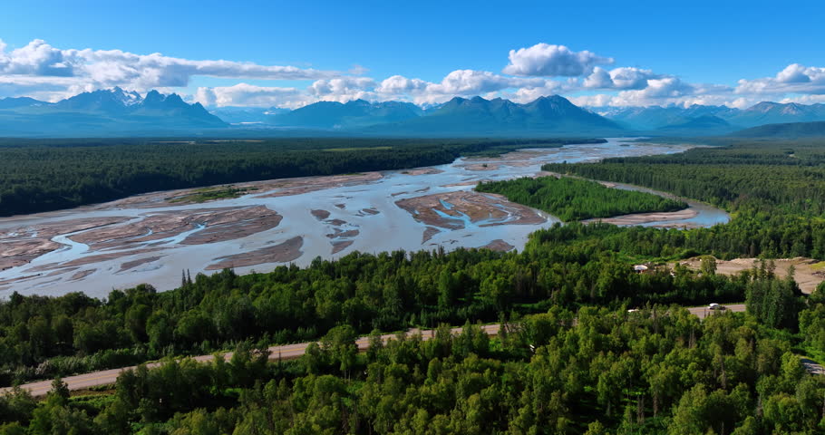 Vast green woods surrounded by the spectacular mountains. Shallow river crosses the pine tree forest. Alaska nature. Aerial view.