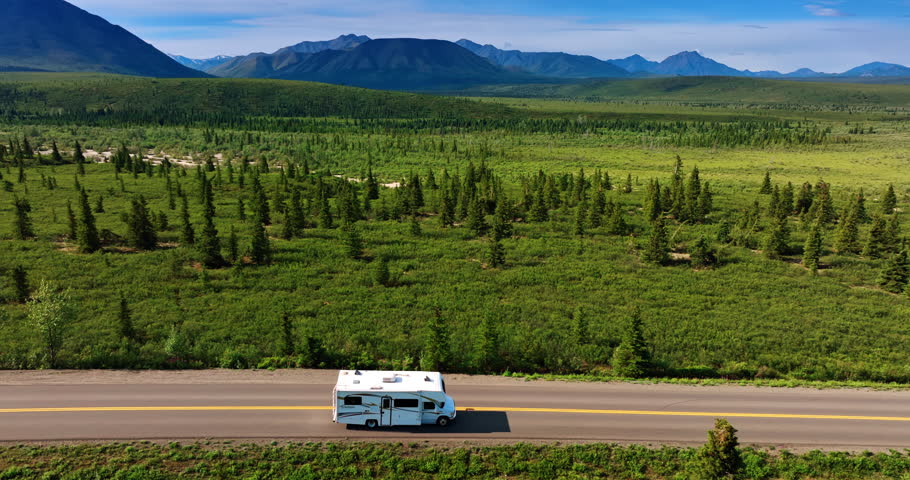 Following a white campervan moving by road in the wilderness. Mountain silhouettes at backdrop. Travel by Alaska.