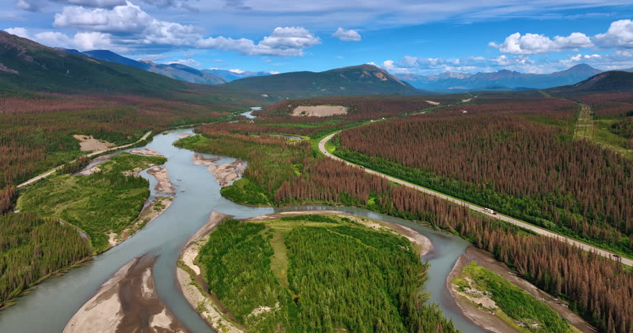 Branching river flowing among the pine tree woods. Few cars move by the highway along the river. Beautiful mountains at backdrop. Alaska.