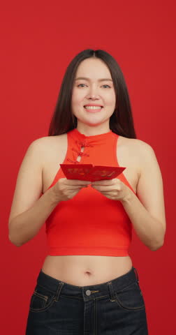 Vertical clip. Happy young Asian woman in red Chinese dress holding red envelopes (Ang Pao) and smiling on red background for Chinese New Year celebration.