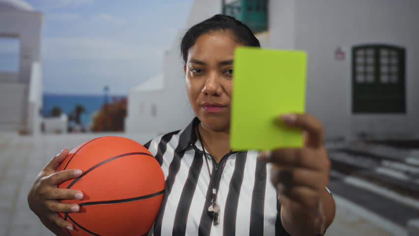 Woman referee holding basketball and showing yellow card while wearing whistle on street; fairness authority.