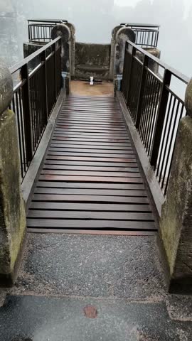 Smooth camera movement from misty Bastei Bridge wooden walkway with metal railings glides toward surrounding Saxon Switzerland mountains, forests, and foggy landscape in Germany.