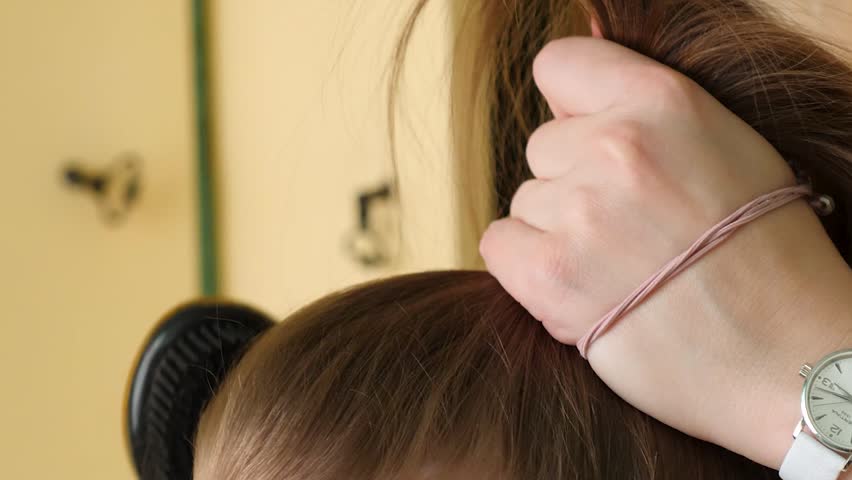 Female hands with pink manicure carefully combing long brown hair of young girl using gold glitter hairbrush to create high ponytail hairstyle.