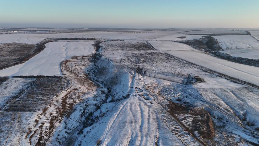 Aerial view of snow-covered landscape with rolling hills, fields, and trees under clear blue sky, for themes about winter countryside, seasonal weather, rural life and agriculture