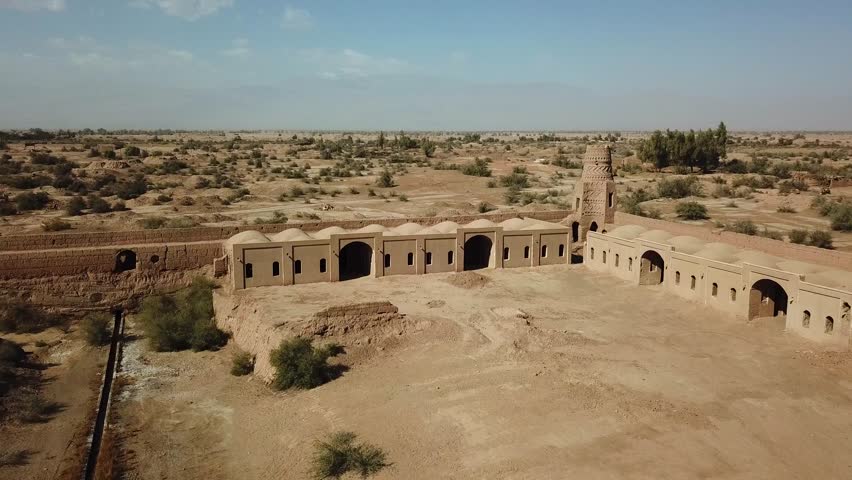 A drone explores ancient ruins situated in a desert. The structures show signs of age and weathering. Open land surrounds the area with sparse vegetation. The sun shines brightly overhead.