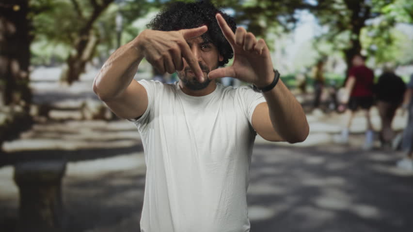 Man framing scene with hands and fingers on street wearing white t shirt and watch; playful curiosity.