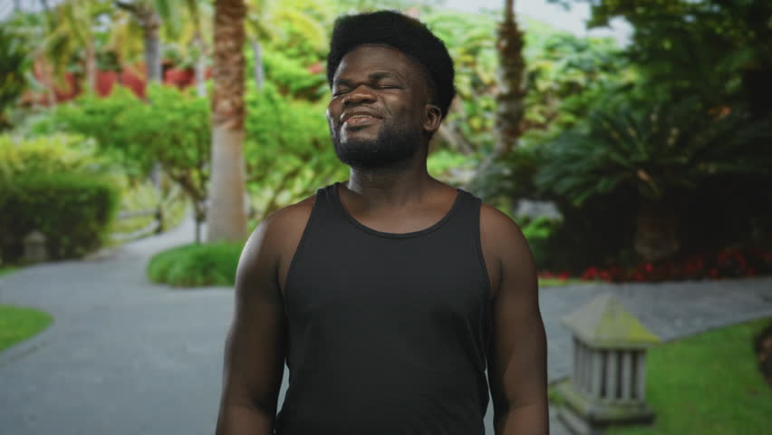 Man with bare shoulders and visible arms tilts head back eyes closed on park path wearing black tank top; calm relief.