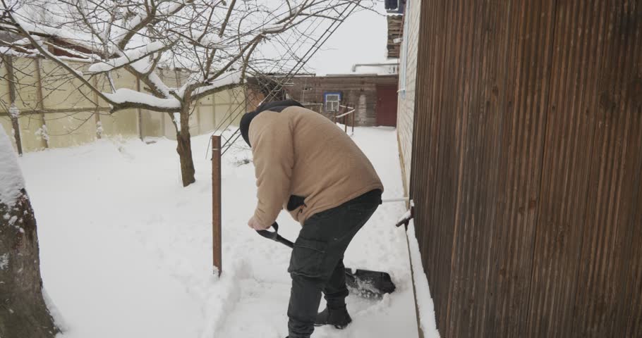 High-angle view of a hardworking man in winter clothing clearing a deep layer of fresh snow from a pathway in his backyard using a shovel, making the walkway safe and accessible after a snowfall