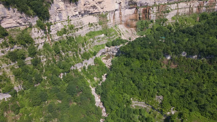 Aerial view shows banded limestone cliff, tiered waterfall, rocky pool, braided stream, and circular metal observation platform in Svaneti, Georgia at midday