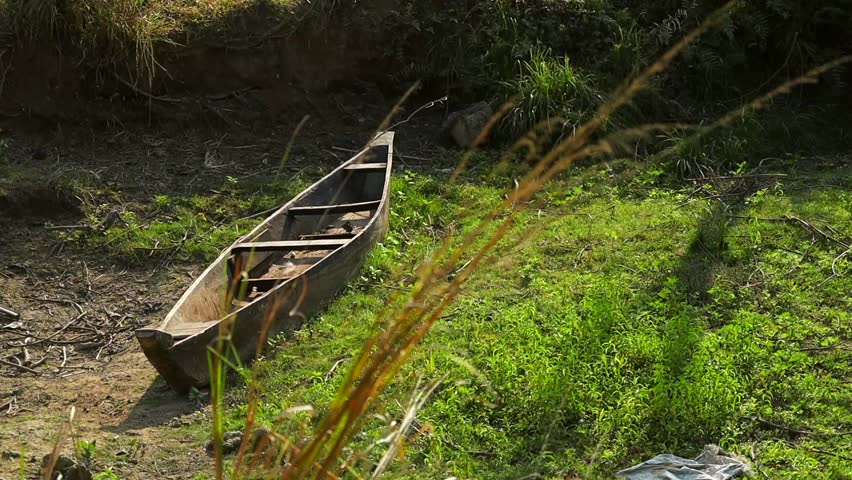 An old wooden canoe lies abandoned on a grassy riverbank, surrounded by wild vegetation and natural textures. Soft sunlight highlights the rustic details of the boat, creating a calm rural atmosphere that reflects simplicity, solitude, and traditional life near water. Ideal for themes related to nature, rural lifestyle, sustainability, travel, heritage, and peaceful outdoor environments.