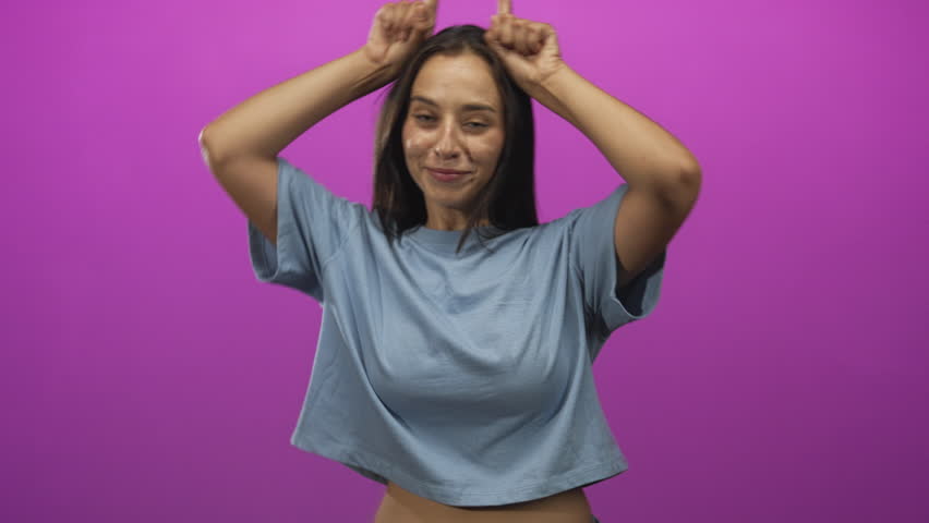 Woman making horn gesture with fingers and smiling, wearing blue crop t shirt in magenta studio; playful confidence.