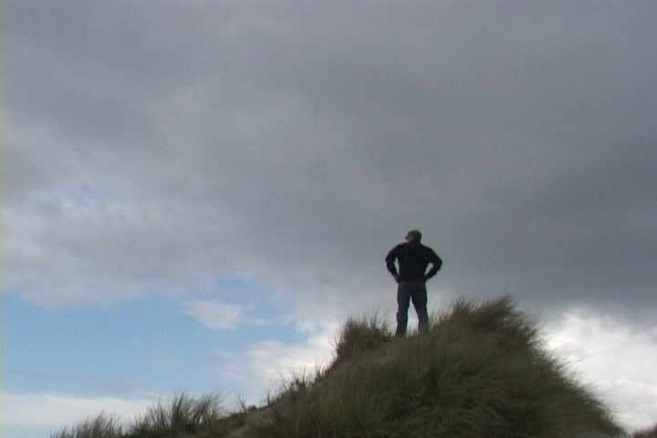 Man walks up sand dune and looks out at view.