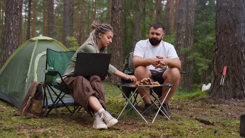 Young couple camping in the woods, with the woman working as a digital nomad on her laptop while the man relaxes, drinks beer, and cooks meat and vegetables on a portable barbecue grill