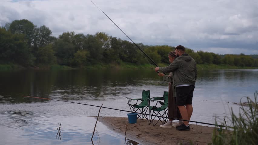 Young couple enjoying a quiet day fishing together, with man patiently teaching woman to cast a fishing rod on the sandy shore of a calm river
