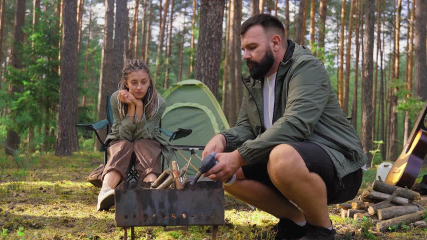 Young couple spending their weekend camping in the pine forest, with the bearded man lighting a fire in the barbecue grill with a gas burner while his girlfriend watches him
