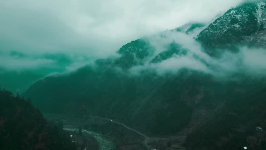 Dramatic clouds swirl around rugged peaks, obscuring a remote valley with a river and road below in the scenic region of Kashmir