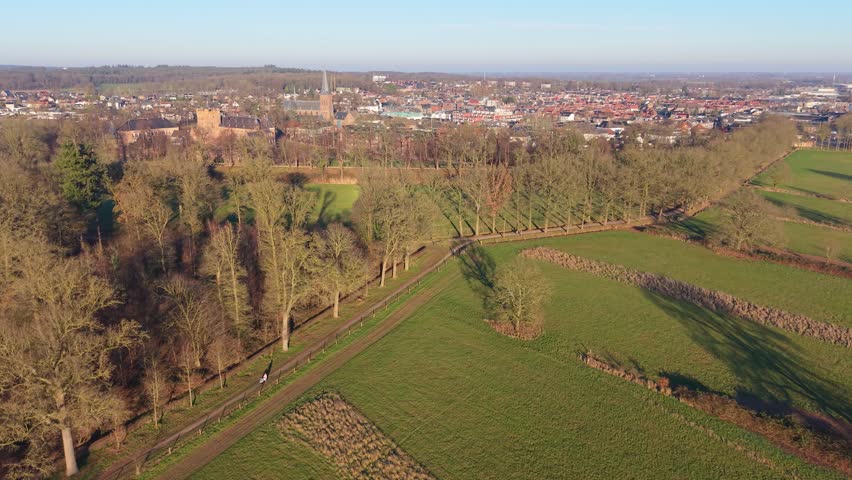Sunlit aerial view of tree lined fields and winding paths leading to a town with church steeple and castle, blending rural geometry with historic architecture.