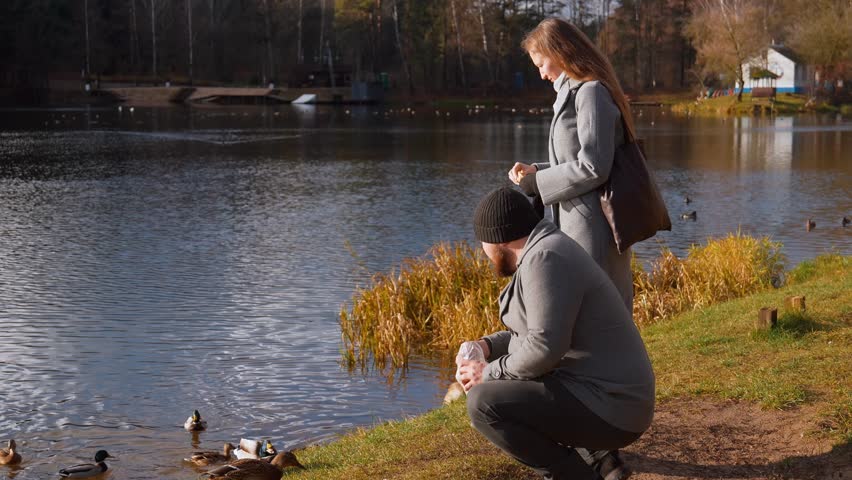 A young couple in love enjoying a sunny autumn day while feeding wild ducks bread swimming in a picturesque lake surrounded by forest, creating a calm and romantic atmosphere of outdoor activities