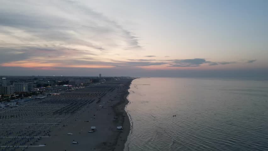 Majestic aerial view flying over a sandy beach during a beautiful sunset. The calm ocean waters meet the shoreline, and city lights twinkle in the background