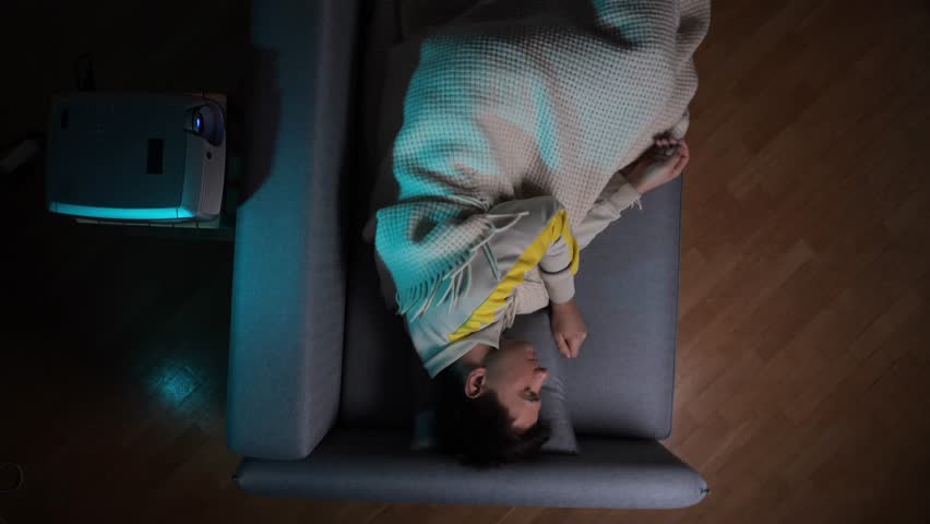 Man lies under a blanket on the sofa, relaxing at home in a dimly lit room projecting a movie, experiencing comfort, leisure, and modern entertainment