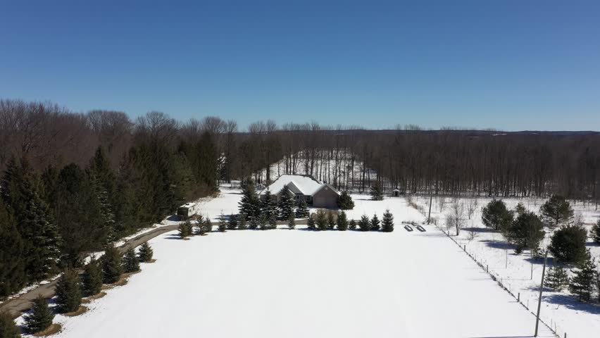 High angle aerial view of a secluded house in the countryside, surrounded by a winter landscape with snow covered ground and bare trees under a clear blue sky on a sunny day