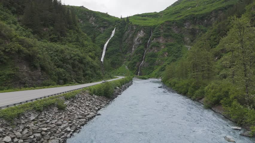 Aerial view over a river in a canyon following a car driving up a road towards a large waterfall.