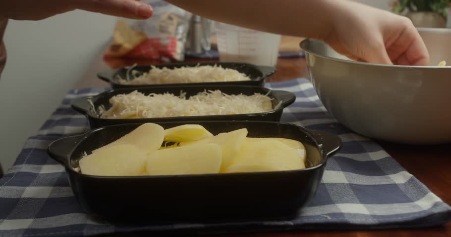 Chef places thinly sliced potatoes into cast iron baking dishes, arranging potato slices like chips. Close-up of homemade cooking process, natural light, traditional comfort food preparation.