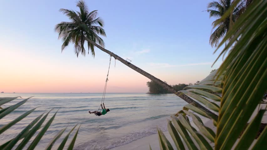 Enjoy the serene beauty of Koh Kood, Thailand, as a woman swings from a palm tree at twilight. The tranquil ocean waves and vibrant hues capture the magic of this Thai paradise.