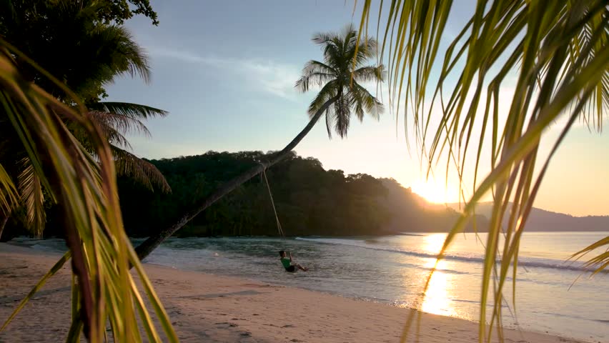 Experience the serene charm of Koh Kood, Thailand, as the sun sets over tranquil waters. Soft waves lap the shore while palm leaves frame the stunning view, a woman on a swing under a palm tree