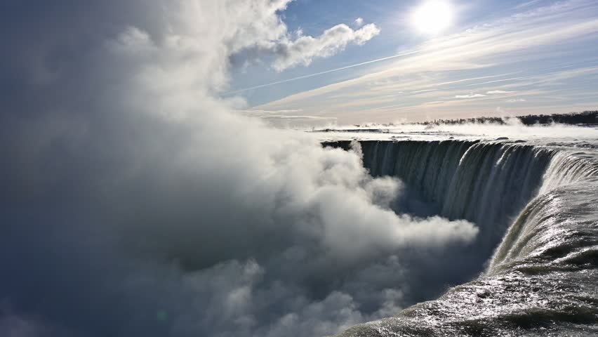 Dramatic winter view of Niagara Falls with thick icy mist and powerful water flow under bright sunlight and clouds. Scenic natural landmark in Ontario, Canada.