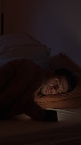 Man lying in bed holding smartphone and looking at screen in night. Concept of insomnia, sleep problems, night phone use and digital addiction