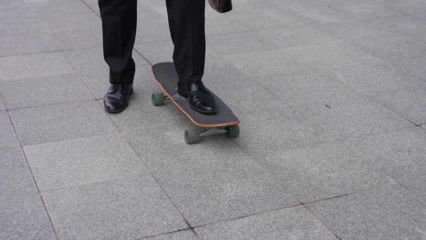 Tilt up shot from dress shoes on skateboard to confident face of Caucasian businessman in dark suit holding brown briefcase skateboarding on paved plaza with glass buildings in Seoul slow motion