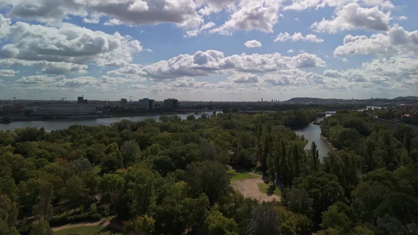 Ascending aerial view of Budapest, Hungary from the Óbudai-sziget during autumn, showcasing Margaret Island along the River Danube, with cityscape, bridges, and urban greenery under a blue cloudy sky.