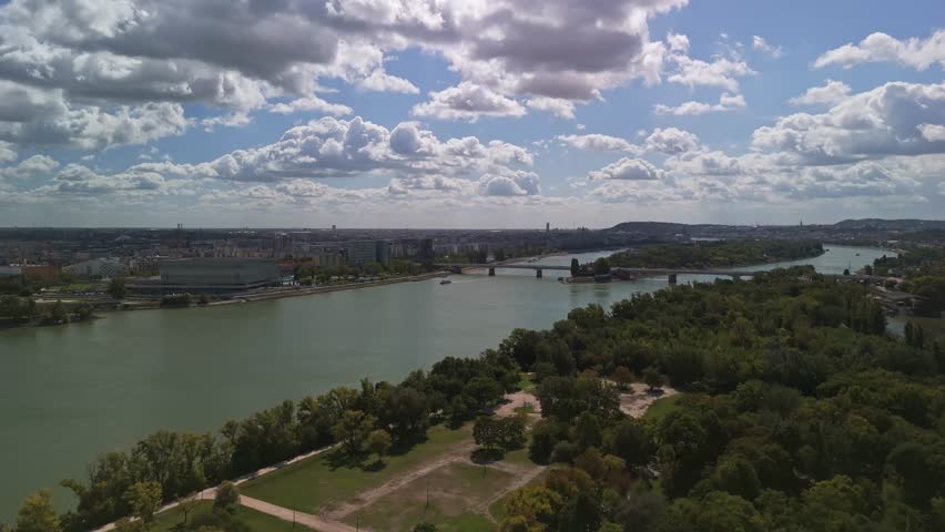 Orbital drone view of Budapest from Hajógyári-sziget, with the view of Margaret Island along the Danube, revealing the cityscape, bridges, and urban greenery under a blue cloudy autumn sky in Hungary.