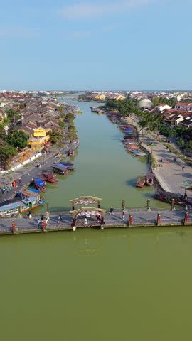 Aerial view of bridge crossing the river in Hoi An old town, with people walking along the waterfront, traditional buildings, and calm water reflecting one of Vietnam