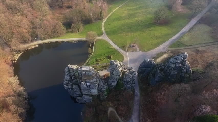 Drone descends toward Externsteine rocks with autumn forest and pond below