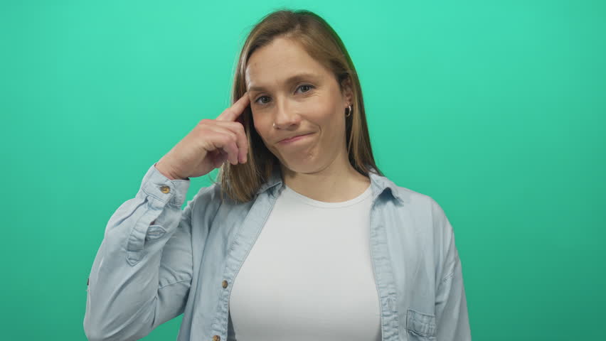 Woman thinking with hand on temple against an isolated green wall, conveying a thoughtful expression in casual attire, blonde hair, youthful demeanor, and casual style.