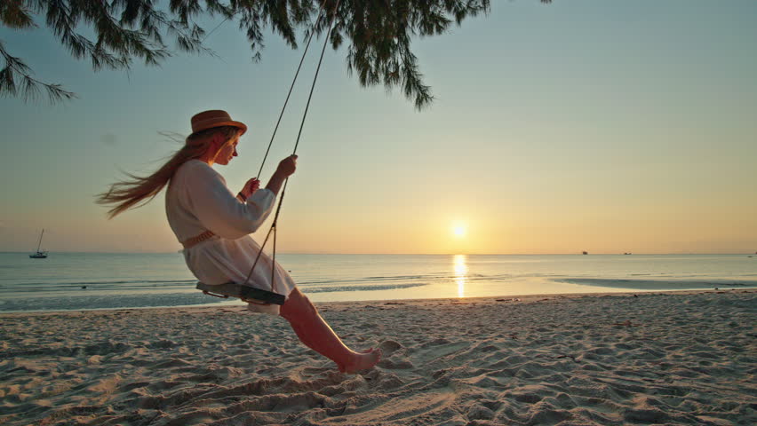 Teenage girl swinging on wooden beach swing at Lipa Noi during sunset and sunrise, silhouette over calm sea with golden reflection, tropical shoreline and peaceful evening mood. High quality 4k