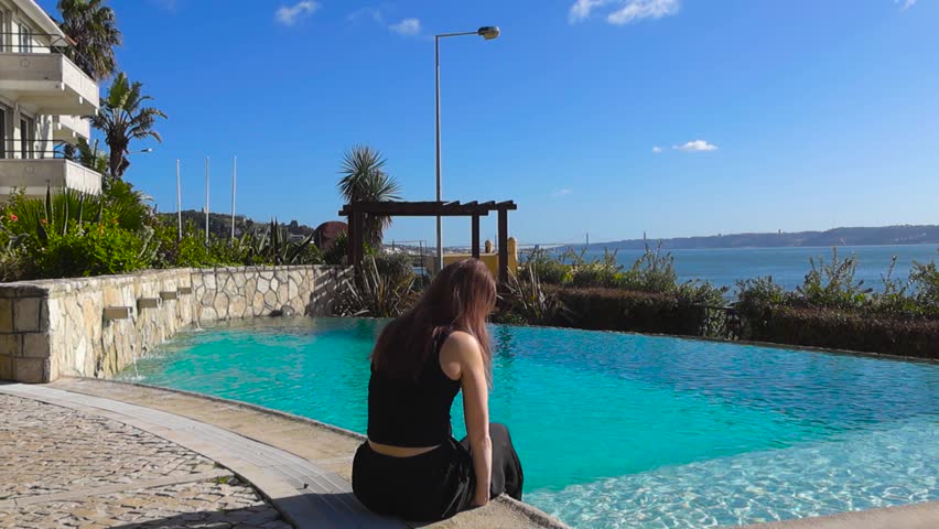 Red-haired woman sits by a luxury hotel pool, dipping feet in turquoise water with sunny Atlantic coastline views in Portugal.