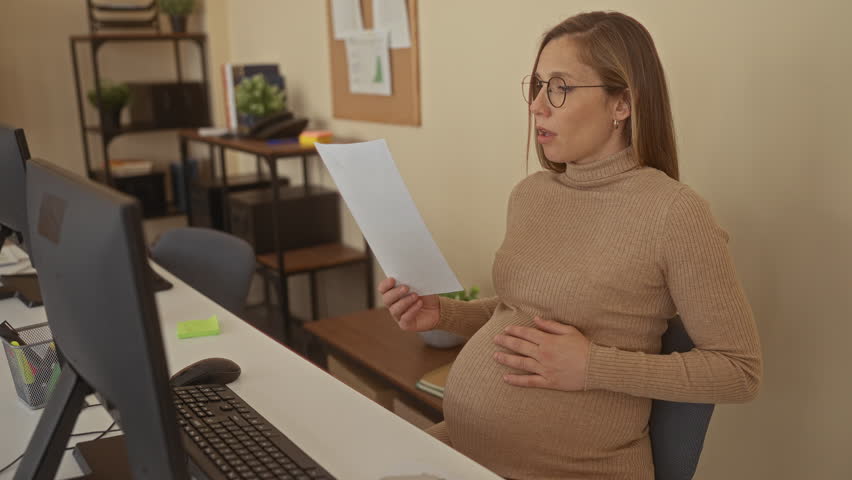 Pregnant woman sitting in office holding document, working at desk, surrounded by computers, emphasizing workplace environment.