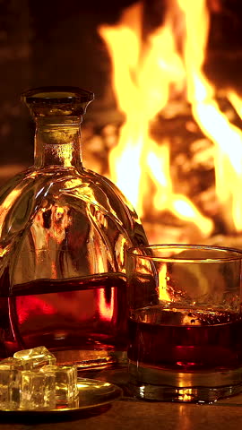 Whiskey glass and bottle near fireplace with warm evening atmosphere, Close up of amber alcohol drink with ice reflecting flames in cozy interior, vertical footage