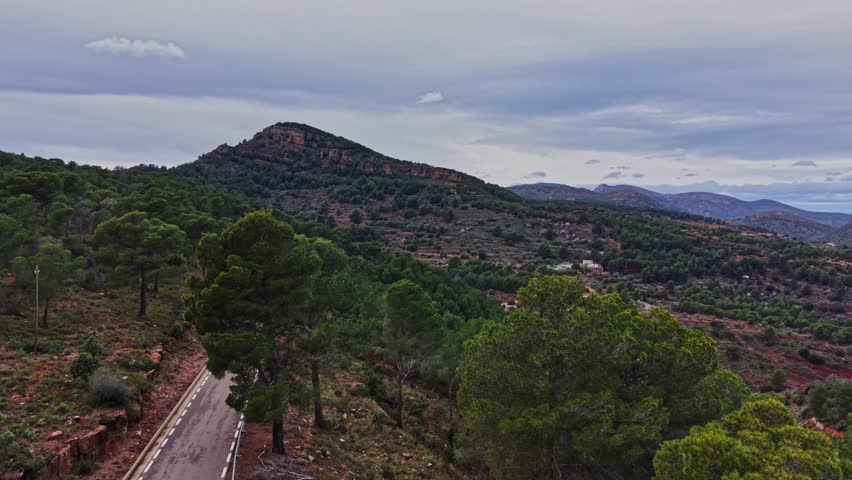 The view shows a mountain landscape in Spain. A winding road cuts through green trees and hills under a cloudy sky. The scene captures nature and the road from above.