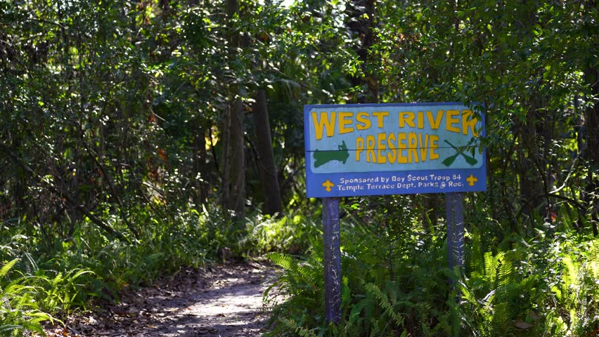 Large blue “West River Preserve” park sign in Temple Terrace, Florida, featuring a canoer graphic and paddles, backed by dense trees, foliage, and natural greenery on a sunny day.