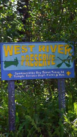 A vertical view of the bright blue West River Preserve sign in Temple Terrace, framed by tall trees and dense foliage, highlighting access to protected wetlands and paddling trails near Tampa.
