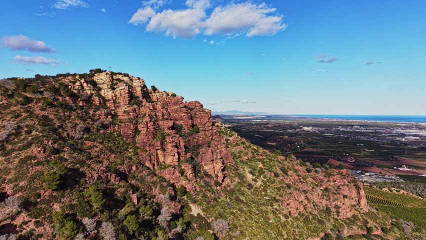 The scene shows rocky hills rising above green fields. The coast is visible in the distance under a clear blue sky. The sunlight highlights the unique rock formations and land below.
