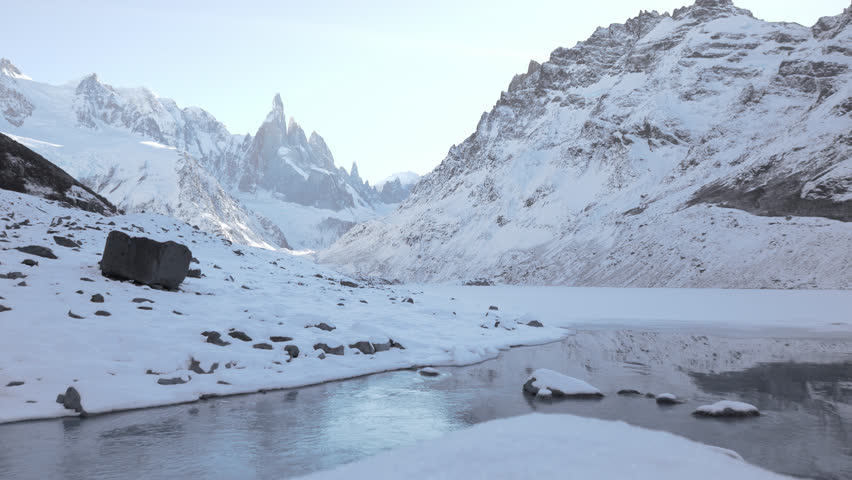 Static shot of Cerro Torre massif rising above Laguna Torre, featuring flowing icy water, snow covered slopes, and dramatic Patagonian peaks.