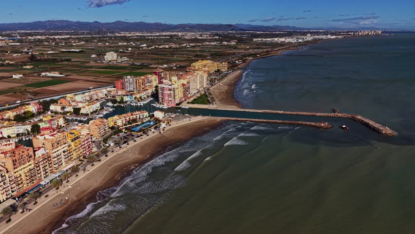 People enjoy the sunny beach while others walk along the pier. Colorful buildings stand near the water. The landscape has fields and mountains in the background.