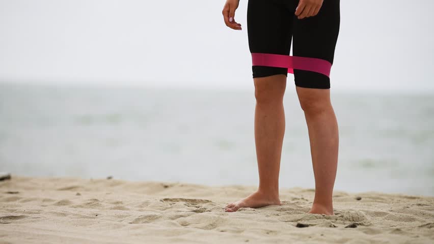 Bare feet performing leg squats and lateral steps with pink resistance band around black shorts on sandy beach; ocean waves and cloudy sky background
